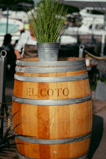 A wooden barrel with the text 'EL COTO' engraved on it, topped with a metal bucket containing tall green grass. The background includes a marina with several blurred boats and a person wearing a white shirt and apron, suggesting a waterfront location.