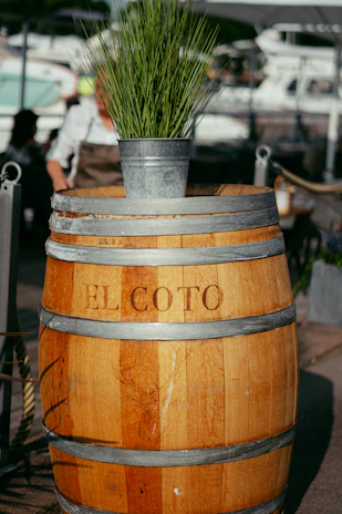 A wooden barrel with the text 'EL COTO' engraved on it, topped with a metal bucket containing tall green grass. The background includes a marina with several blurred boats and a person wearing a white shirt and apron, suggesting a waterfront location.