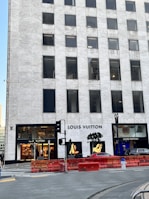 A tall, marble-clad building with the Louis Vuitton store prominently on the ground floor. The storefront is framed by large glass windows showcasing high-end merchandise and displays. A couple of trees are positioned near the entrance, and there are some red barriers in front of the building. A luxurious car is partially visible nearby.