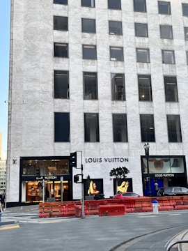 A tall, marble-clad building with the Louis Vuitton store prominently on the ground floor. The storefront is framed by large glass windows showcasing high-end merchandise and displays. A couple of trees are positioned near the entrance, and there are some red barriers in front of the building. A luxurious car is partially visible nearby.
