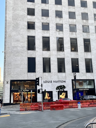 A tall, marble-clad building with the Louis Vuitton store prominently on the ground floor. The storefront is framed by large glass windows showcasing high-end merchandise and displays. A couple of trees are positioned near the entrance, and there are some red barriers in front of the building. A luxurious car is partially visible nearby.