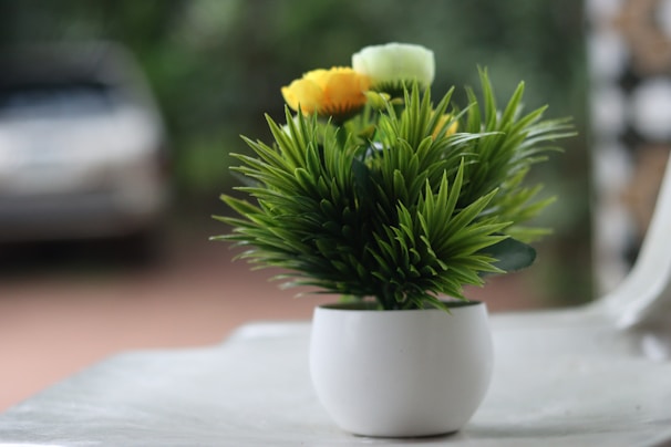 A small white pot contains a dense arrangement of green artificial grass-like plants with yellow and light green artificial flowers. The pot is placed on a smooth, light-colored surface, with a blurred background that suggests an outdoor setting.