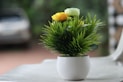 A small white pot contains a dense arrangement of green artificial grass-like plants with yellow and light green artificial flowers. The pot is placed on a smooth, light-colored surface, with a blurred background that suggests an outdoor setting.