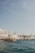 A coastal scene features several buildings with a Mediterranean architectural style, situated along the shoreline. People are visible on the rocky edge overlooking the calm, blue sea. The sky is clear with a few wispy clouds.