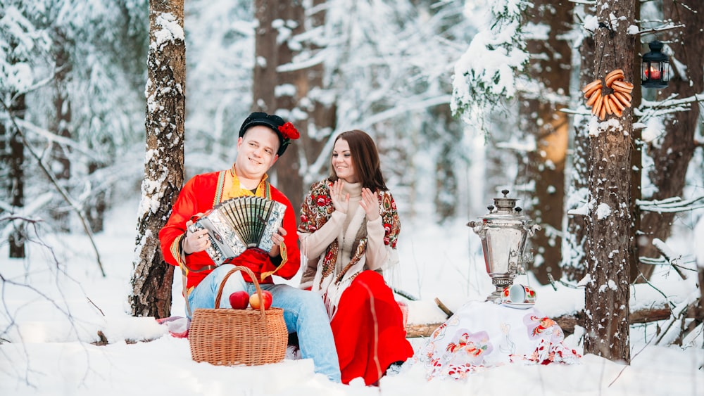 a man and woman sitting in the snow with a basket of apples