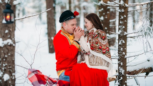 A couple dressed in traditional attire shares an intimate moment in a snowy forest setting. The man wears a red shirt with gold embroidery and a black hat, while the woman is adorned with a colorful shawl over her shoulders. They are holding hands, sitting on a blanket-covered bench, surrounded by snow-covered trees. A lantern hangs nearby, adding warmth to the winter scene.