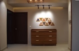 A modern interior hallway features a wooden dresser with three drawers against a light-colored wall. Nine triangular art pieces in various colors and patterns are arranged in a grid above the dresser. The ceiling has track lighting focused on the artwork, and there's a dark wooden door to the left.