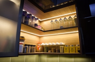 A well-organized kitchen shelf featuring a variety of containers and jars in different sizes. The top shelf holds transparent containers with dry goods. The middle shelf showcases jars with metallic lids, some filled with spices or seeds. The lower shelf contains labeled containers for tea, sugar, and coffee in bright yellow with black text. The lighting creates a warm, inviting ambience.