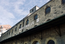 An industrial building with stone brick walls, featuring arched windows and a large sign reading 'WERFTHALLE III'. The roof structure is visible along with some architectural details. The sky is partially cloudy, adding a textured background.
