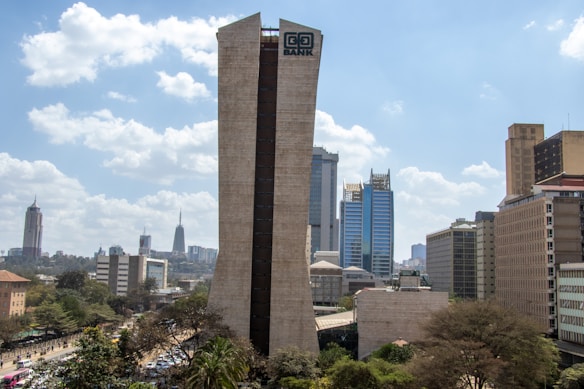 A cityscape featuring a prominent tall building marked with a bank's name. Surrounding the central skyscraper are other modern and distinctive high-rise structures. The sky is filled with scattered clouds, and there are trees and a busy road at street level.