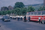 A marching band dressed in uniforms is walking along a street beside a vintage red bus. The scene includes several people walking nearby and an old black car traveling on the road. The background features trees and historic stone buildings.