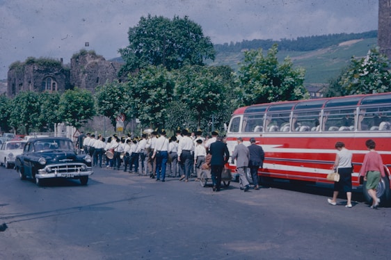 A marching band dressed in uniforms is walking along a street beside a vintage red bus. The scene includes several people walking nearby and an old black car traveling on the road. The background features trees and historic stone buildings.