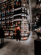 A modern bookstore with an extensive collection of books lining the tall, sleek shelves. The lighting is warm and inviting, creating a cozy atmosphere. Two individuals can be seen, one wearing a backpack and standing near the shelves, and another browsing books further down the aisle. The floor is tiled with large, dark gray tiles, complementing the sophisticated interior design.