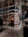 A modern bookstore with an extensive collection of books lining the tall, sleek shelves. The lighting is warm and inviting, creating a cozy atmosphere. Two individuals can be seen, one wearing a backpack and standing near the shelves, and another browsing books further down the aisle. The floor is tiled with large, dark gray tiles, complementing the sophisticated interior design.