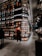 A modern bookstore with an extensive collection of books lining the tall, sleek shelves. The lighting is warm and inviting, creating a cozy atmosphere. Two individuals can be seen, one wearing a backpack and standing near the shelves, and another browsing books further down the aisle. The floor is tiled with large, dark gray tiles, complementing the sophisticated interior design.