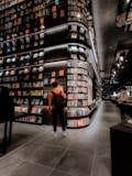 A modern bookstore with an extensive collection of books lining the tall, sleek shelves. The lighting is warm and inviting, creating a cozy atmosphere. Two individuals can be seen, one wearing a backpack and standing near the shelves, and another browsing books further down the aisle. The floor is tiled with large, dark gray tiles, complementing the sophisticated interior design.