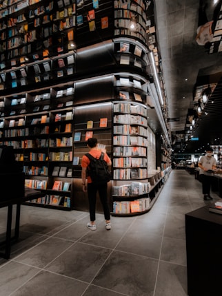 A modern bookstore with an extensive collection of books lining the tall, sleek shelves. The lighting is warm and inviting, creating a cozy atmosphere. Two individuals can be seen, one wearing a backpack and standing near the shelves, and another browsing books further down the aisle. The floor is tiled with large, dark gray tiles, complementing the sophisticated interior design.