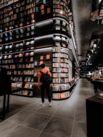 A modern bookstore with an extensive collection of books lining the tall, sleek shelves. The lighting is warm and inviting, creating a cozy atmosphere. Two individuals can be seen, one wearing a backpack and standing near the shelves, and another browsing books further down the aisle. The floor is tiled with large, dark gray tiles, complementing the sophisticated interior design.