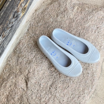 A pair of white rubber shoes placed on a textured stone surface. The stone has a rough, natural appearance with hues of beige and light brown. The shoes are simple in design with some printed text inside, and they are positioned parallel to each other. Wooden planks can be seen partially in the background, suggesting an outdoor setting.