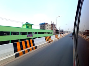 A highway or expressway with clear markings and an extended view of a cityscape. To the left, a bright green building and an under-construction structure are visible. The road is flanked by black and orange striped barriers, and there is a reflection of another vehicle with a person’s arm visible.