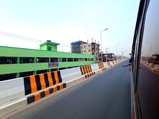 A highway or expressway with clear markings and an extended view of a cityscape. To the left, a bright green building and an under-construction structure are visible. The road is flanked by black and orange striped barriers, and there is a reflection of another vehicle with a person’s arm visible.