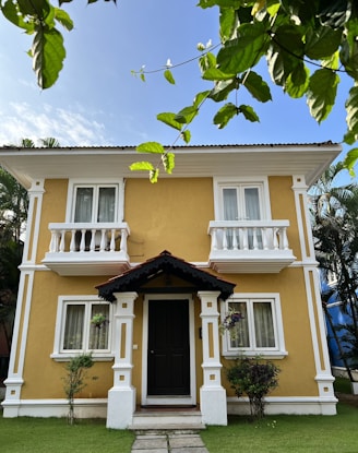 A two-story house with a warm yellow exterior and white decorative trim. It features small balconies, a central front door with a dark color, and a small porch. Greenery surrounds the house with plants, trees, and a well-manicured lawn. The sky is clear and blue, and there are tree branches with green leaves in the foreground.