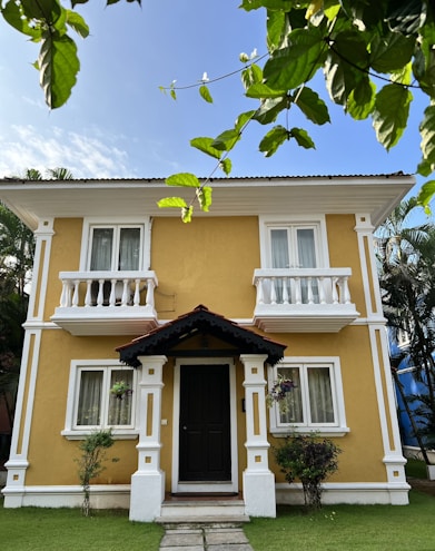 A two-story house with a warm yellow exterior and white decorative trim. It features small balconies, a central front door with a dark color, and a small porch. Greenery surrounds the house with plants, trees, and a well-manicured lawn. The sky is clear and blue, and there are tree branches with green leaves in the foreground.