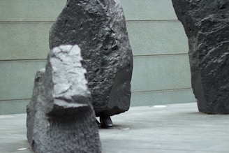 Large, rough-textured stone sculptures are positioned on a smooth, grey floor. The background features a wall with horizontal lines. Part of a person's shoe is visible peeking out from behind one of the stones.