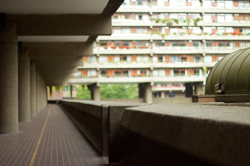 A long, concrete corridor on the left with multiple support beams leading to a large, modern residential building in the background. The structure has a brutalist architectural style, characterized by rough, unfinished surfaces and repetitive modular elements. Greenery and trees are visible, and there is a dome-shaped structure on the right.