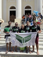 A group of five people standing on steps in front of a building with tall, white columns. They are holding a banner with the word 'LUMUS' and some are wearing matching black t-shirts. In the background, there are more people, some of whom are seated or standing around, and a few are holding inflatable toys.