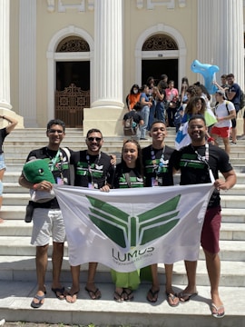 A group of five people standing on steps in front of a building with tall, white columns. They are holding a banner with the word 'LUMUS' and some are wearing matching black t-shirts. In the background, there are more people, some of whom are seated or standing around, and a few are holding inflatable toys.