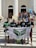A group of five people standing on steps in front of a building with tall, white columns. They are holding a banner with the word 'LUMUS' and some are wearing matching black t-shirts. In the background, there are more people, some of whom are seated or standing around, and a few are holding inflatable toys.