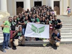 A group of young adults are gathered on the steps of a building with white pillars. They are holding a banner with green insignia. Most are wearing black t-shirts with matching designs. The group appears happy and energetic, posing for the photo with various hand signs.