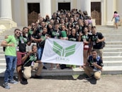 A group of young adults are gathered on the steps of a building with white pillars. They are holding a banner with green insignia. Most are wearing black t-shirts with matching designs. The group appears happy and energetic, posing for the photo with various hand signs.