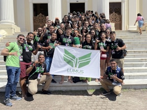 A group of young adults are gathered on the steps of a building with white pillars. They are holding a banner with green insignia. Most are wearing black t-shirts with matching designs. The group appears happy and energetic, posing for the photo with various hand signs.