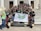 A group of young adults are gathered on the steps of a building with white pillars. They are holding a banner with green insignia. Most are wearing black t-shirts with matching designs. The group appears happy and energetic, posing for the photo with various hand signs.