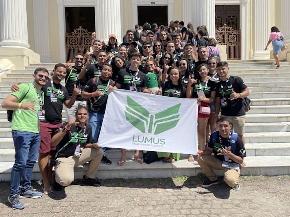 A group of young adults are gathered on the steps of a building with white pillars. They are holding a banner with green insignia. Most are wearing black t-shirts with matching designs. The group appears happy and energetic, posing for the photo with various hand signs.