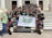 A group of young adults are gathered on the steps of a building with white pillars. They are holding a banner with green insignia. Most are wearing black t-shirts with matching designs. The group appears happy and energetic, posing for the photo with various hand signs.