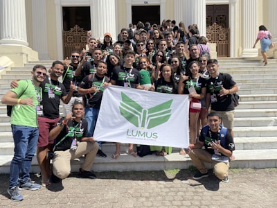 A group of young adults are gathered on the steps of a building with white pillars. They are holding a banner with green insignia. Most are wearing black t-shirts with matching designs. The group appears happy and energetic, posing for the photo with various hand signs.