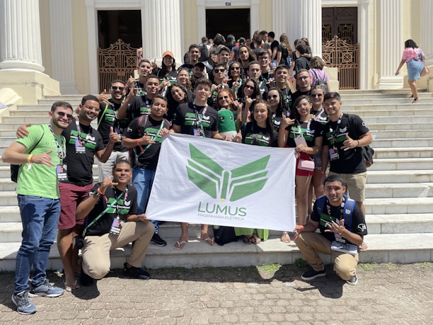 A group of young adults are gathered on the steps of a building with white pillars. They are holding a banner with green insignia. Most are wearing black t-shirts with matching designs. The group appears happy and energetic, posing for the photo with various hand signs.