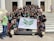 A group of young adults are gathered on the steps of a building with white pillars. They are holding a banner with green insignia. Most are wearing black t-shirts with matching designs. The group appears happy and energetic, posing for the photo with various hand signs.
