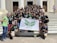 A group of young adults are gathered on the steps of a building with white pillars. They are holding a banner with green insignia. Most are wearing black t-shirts with matching designs. The group appears happy and energetic, posing for the photo with various hand signs.