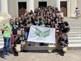 A group of young adults are gathered on the steps of a building with white pillars. They are holding a banner with green insignia. Most are wearing black t-shirts with matching designs. The group appears happy and energetic, posing for the photo with various hand signs.