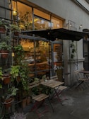A cozy café exterior with lush green plants surrounding the entrance and a small outdoor seating area featuring two wooden tables and folding chairs. The café's large windows reveal a warm, inviting interior with soft lighting. A person is visible inside, suggesting a welcoming atmosphere. The setting is serene and inviting, with greenery adding a natural touch.