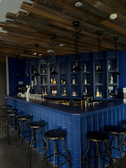 A modern bar area featuring a dark blue tiled counter with several black bar stools arranged around it. The wall behind the counter is also tiled in blue with shelves displaying various bottles of liquor. There are multiple black pendant lights hanging from a wooden, rustic ceiling, creating a cozy and intimate atmosphere. Empty glasses are neatly arranged in a row on the counter, and a small chalkboard menu is visible.