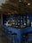 A modern bar area featuring a dark blue tiled counter with several black bar stools arranged around it. The wall behind the counter is also tiled in blue with shelves displaying various bottles of liquor. There are multiple black pendant lights hanging from a wooden, rustic ceiling, creating a cozy and intimate atmosphere. Empty glasses are neatly arranged in a row on the counter, and a small chalkboard menu is visible.