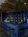 A modern bar area featuring a dark blue tiled counter with several black bar stools arranged around it. The wall behind the counter is also tiled in blue with shelves displaying various bottles of liquor. There are multiple black pendant lights hanging from a wooden, rustic ceiling, creating a cozy and intimate atmosphere. Empty glasses are neatly arranged in a row on the counter, and a small chalkboard menu is visible.