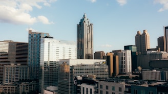 A cityscape featuring a mix of tall modern skyscrapers and older buildings, set against a clear blue sky with a few clouds. The architecture is diverse, with glass and concrete structures dominating the skyline.