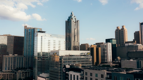 A cityscape featuring a mix of tall modern skyscrapers and older buildings, set against a clear blue sky with a few clouds. The architecture is diverse, with glass and concrete structures dominating the skyline.