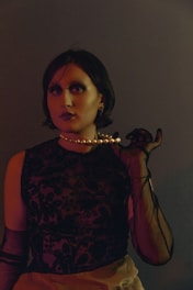A person with short dark hair is wearing a black lace top and sheer gloves, holding a strand of pearls near their neck. The lighting casts dramatic shadows, emphasizing the vintage and mysterious aesthetic.